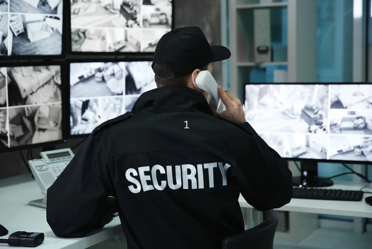 Uniformed security officer in a jacket marked Security speaking on a telephone while watching banks of CCTV monitors